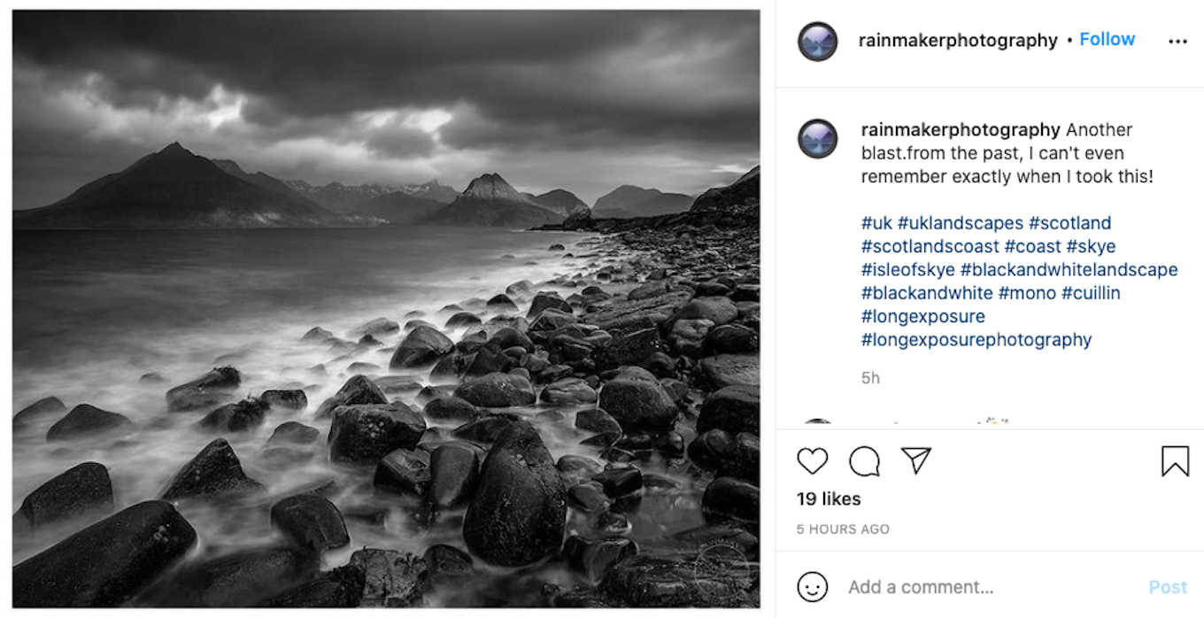 An Instagram screenshot of the ocean in black and white with rocks in the foreground and mountains in the background.